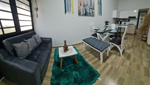 A living room with a grey sofa, a small white table with a statue, and a teal rug. There is a dining area with a white table and chairs, and a kitchen in the background.