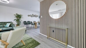 Een moderne woonkamer met een groene bank, een witte fauteuil, een eettafel en een wandpaneel van hout met een ronde spiegel.