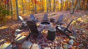 Een boslandschap met een vuurkuil en Adirondack-stoelen eromheen.