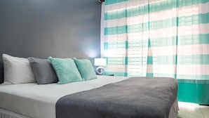 Et soveværelse med en seng, puder, et natbord med en lampe og et vindue med stribede gardiner.