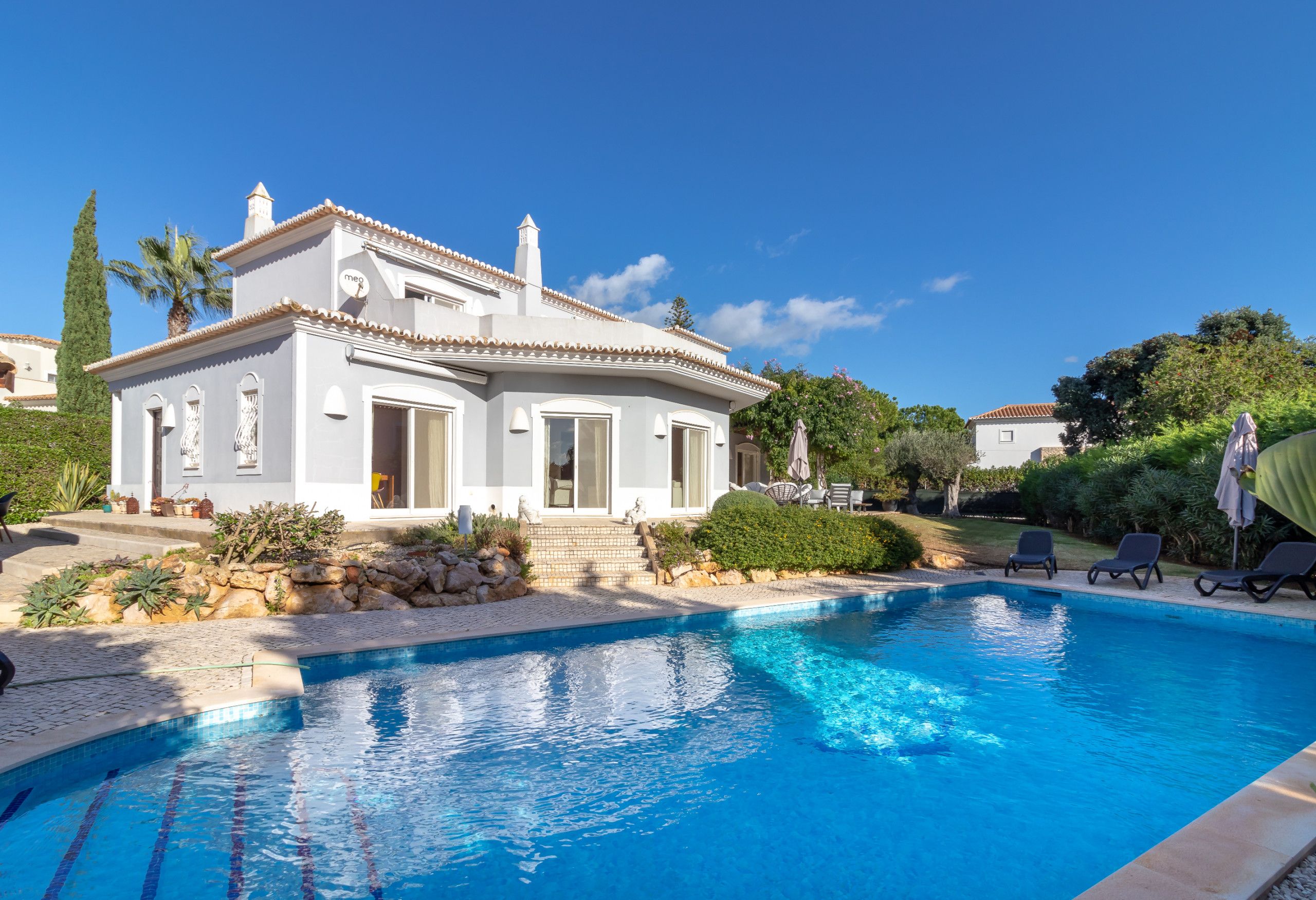 A white villa with a swimming pool, surrounded by greenery and a clear blue sky.