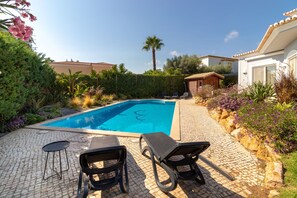 Une piscine entourée de chaises longues et d’une petite table, le tout agrémenté de verdure et bordé d’un chemin en pierre.