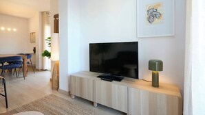 Un salon moderne avec un téléviseur à écran plat posé sur une console en bois, un coin repas avec des chaises bleues et une œuvre d’art encadrée accrochée au mur.