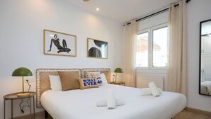 Un lit double bien fait, avec du linge de lit blanc, deux oreillers et une serviette enroulée. On y trouve deux tables de chevet avec des lampes, une fenêtre avec des rideaux et deux œuvres d’art encadrées accrochées au mur.
