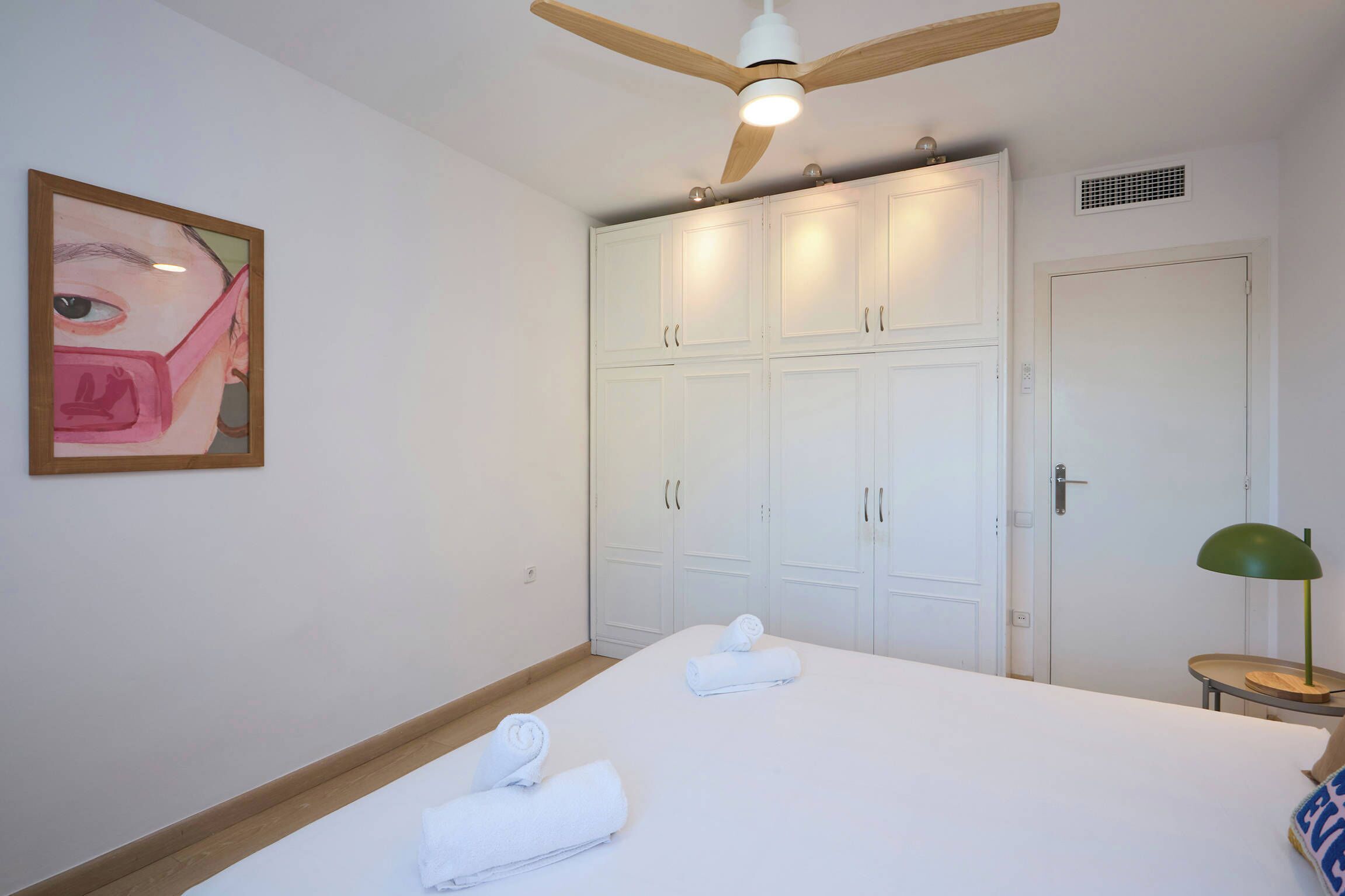 Een hotelkamer met een bed, twee opgerolde handdoeken, een plafondventilator, een schilderij en een lamp.
