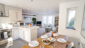 Une cuisine moderne avec des meubles blancs, un coin repas avec une table en bois mise pour le repas, et un espace de vie avec un canapé et un téléviseur.