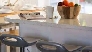 Un comptoir de cuisine avec un saladier en bois rempli de fruits, une tasse blanche et des tabourets de bar.