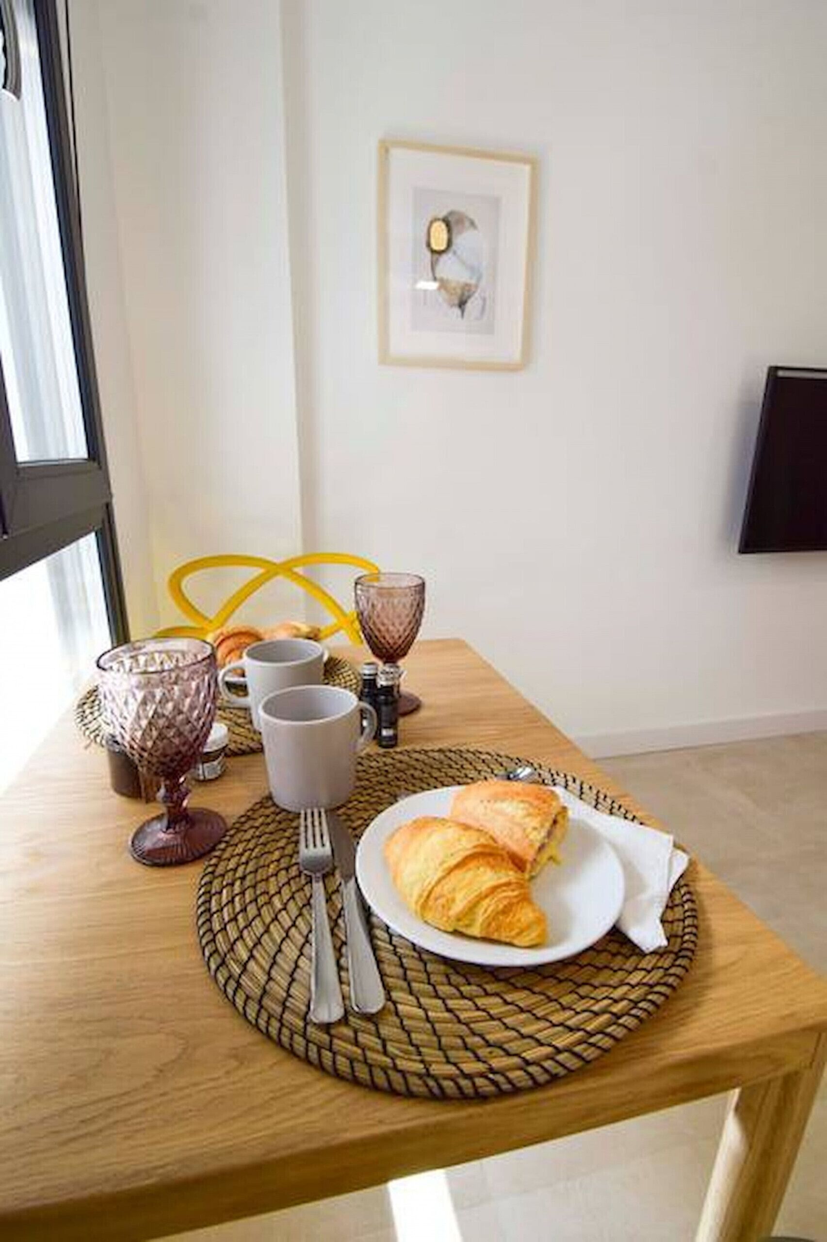 Ett frukostbord med croissanter, en tallrik, bestick och en inramad tavla på väggen.