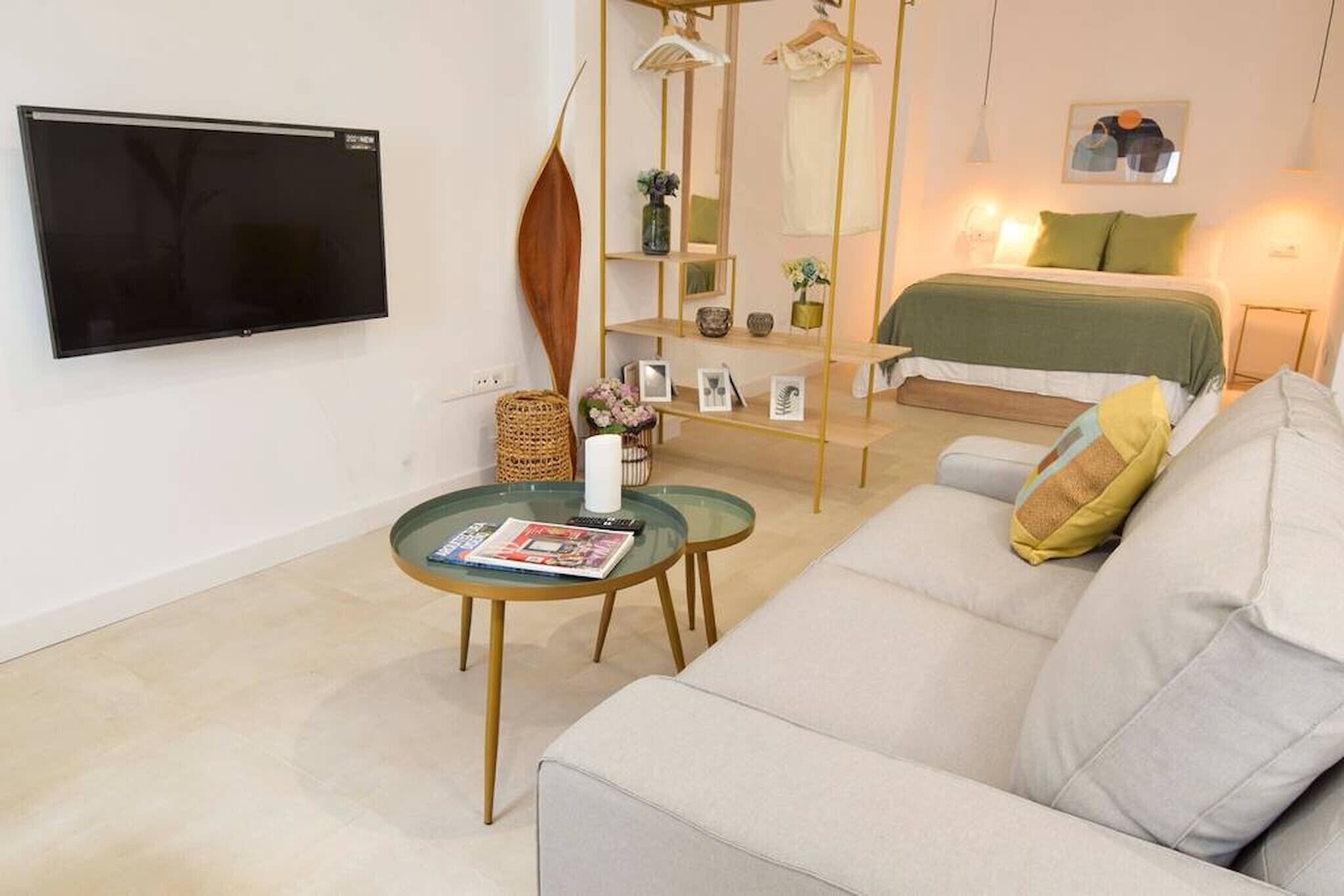 A modern living room with a sofa, a glass coffee table, a TV mounted on the wall, and a bed with a green bedspread.