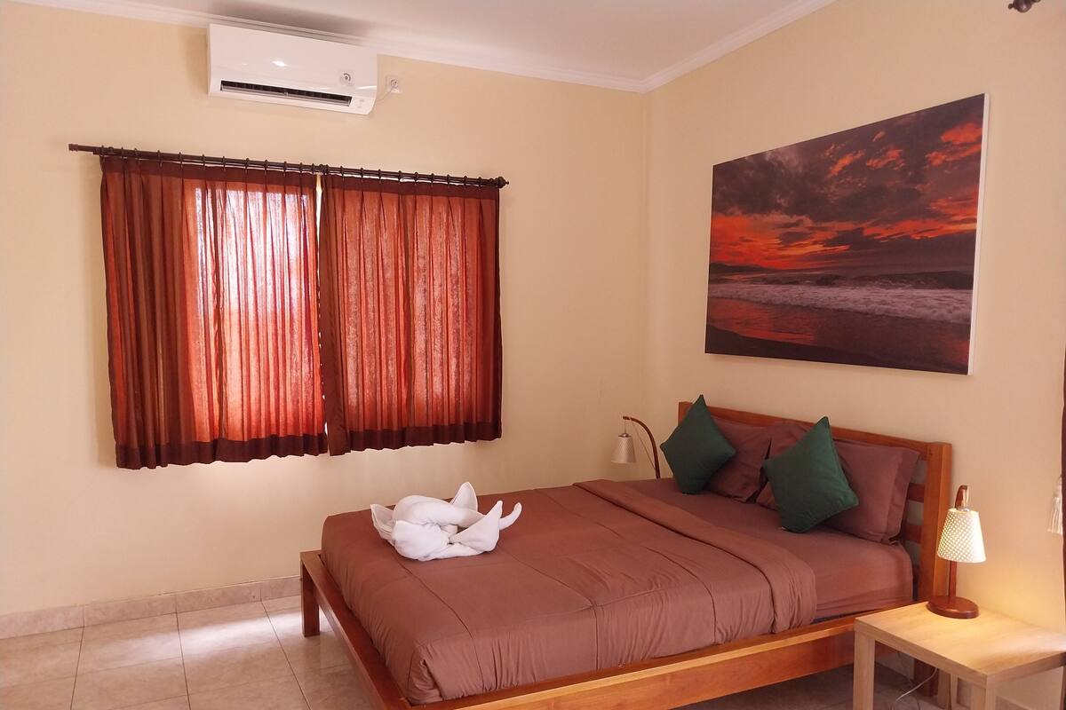 A bedroom with a bed, a nightstand, a lamp, a painting, and a window with curtains.