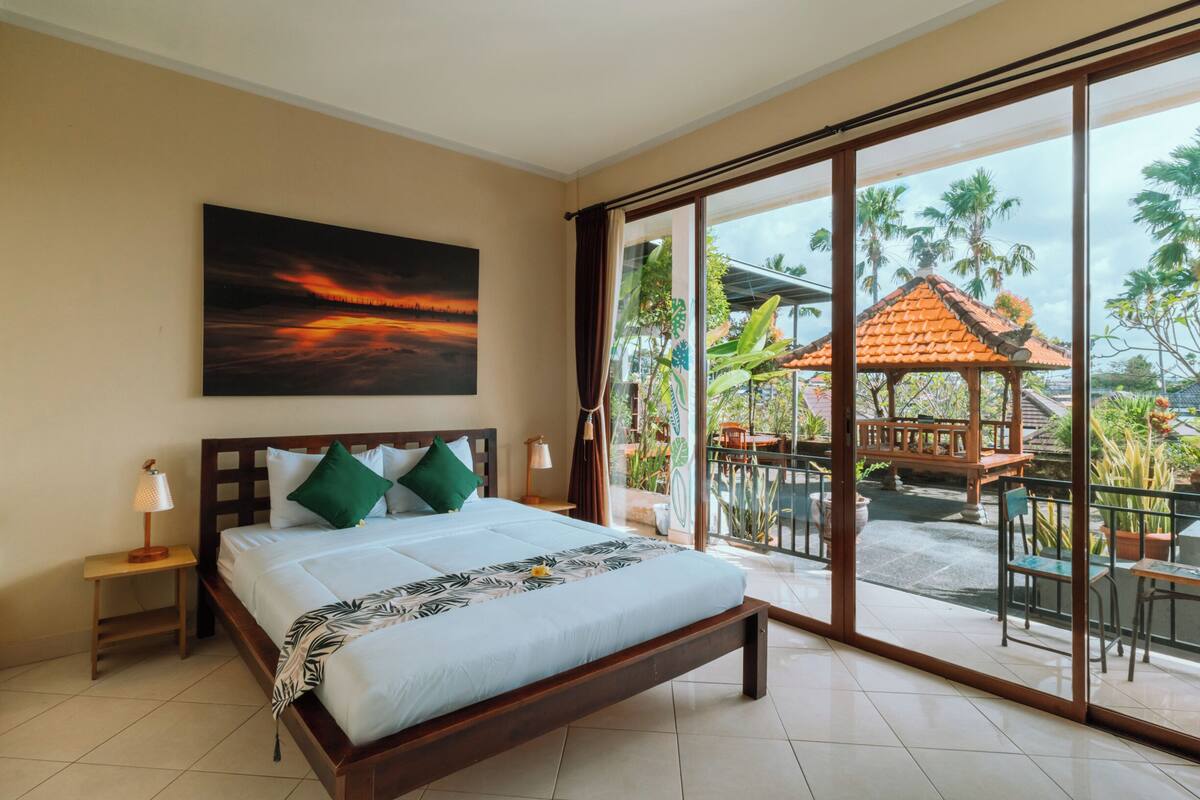 A bedroom with a large bed, a nightstand, a wall-mounted lamp, and a view of a balcony with outdoor furniture and plants.