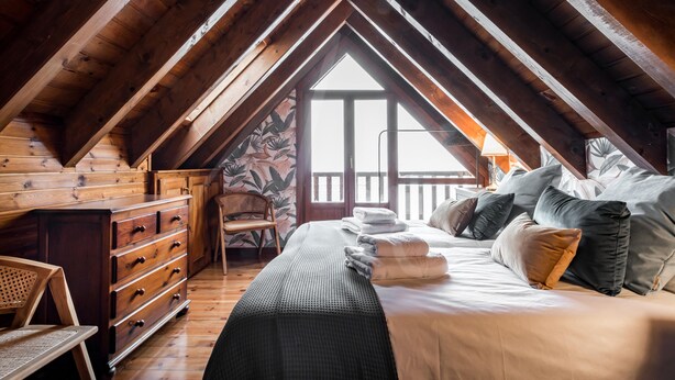 A cozy attic bedroom with a wooden ceiling, a large bed with pillows, a dresser, and a chair. - Tanau 1700 (Tanau)