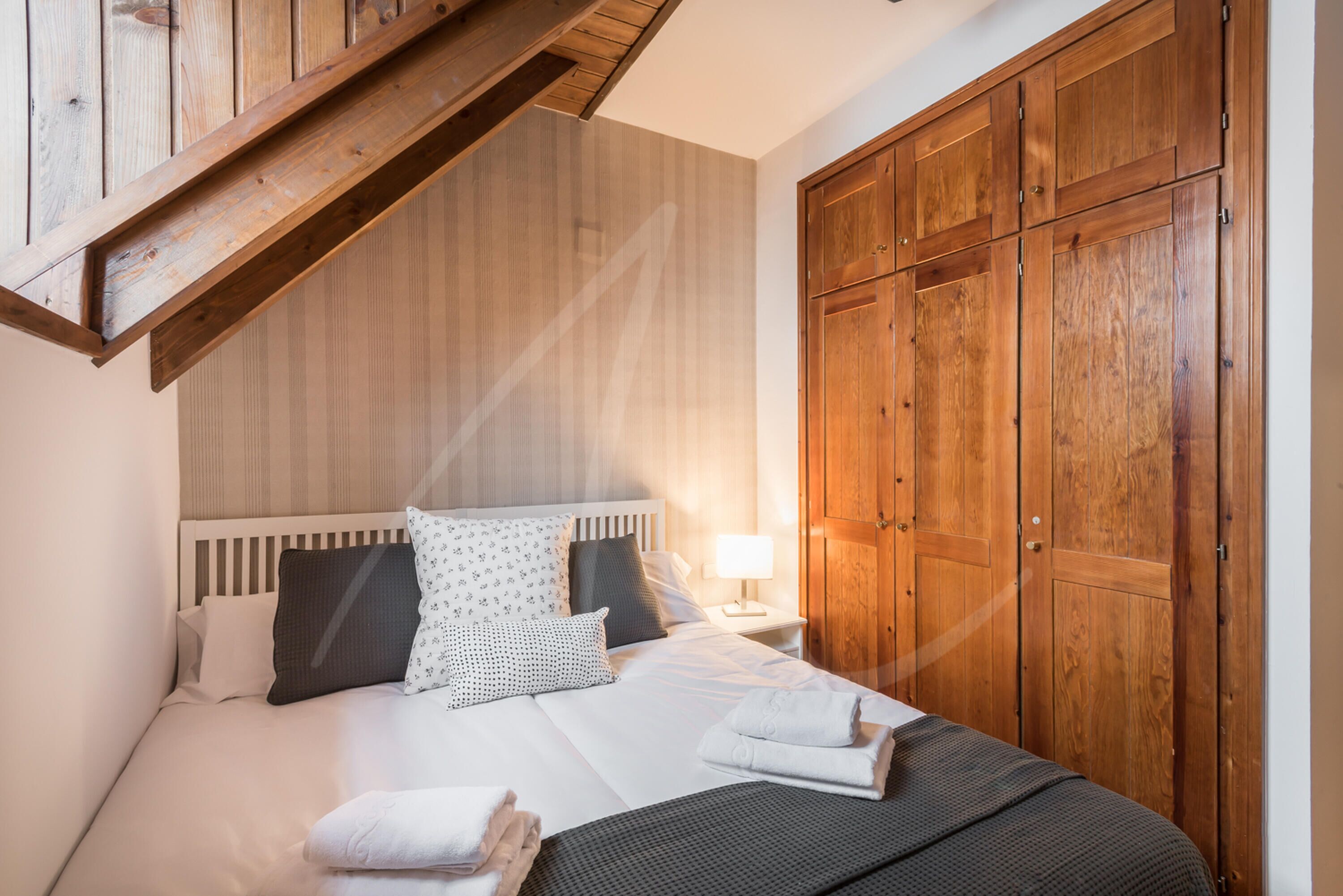 A bedroom with a wooden ceiling, a large wardrobe, a bed with white bedding, and a nightstand with a lamp.
