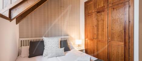 A bedroom with a wooden ceiling, a large wardrobe, a bed with white bedding, and a nightstand with a lamp.