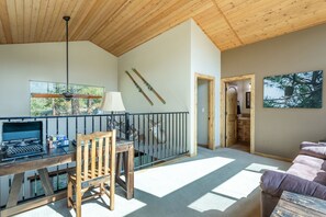 Une pièce spacieuse avec un plafond en bois, un bureau avec un ordinateur, une chaise et un canapé. Des fenêtres laissent entrer la lumière naturelle, et une porte donne accès à une autre pièce.