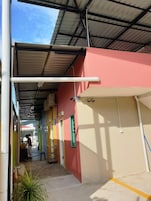 Un espacio al aire libre cubierto con techo metálico, pared roja y pared amarilla con una ventana. Hay una persona trabajando cerca de una ventana.
