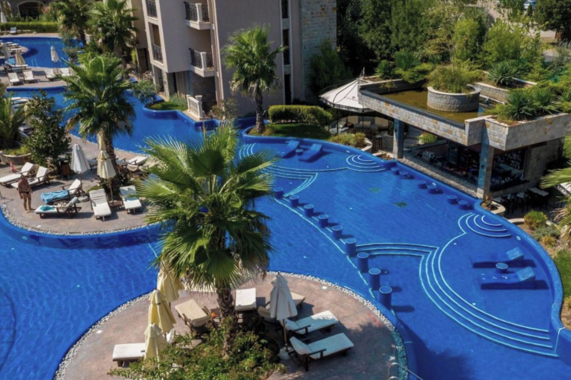 Un área de piscina con sillas de descanso y sombrillas, rodeada de palmeras y un edificio con balcones.
