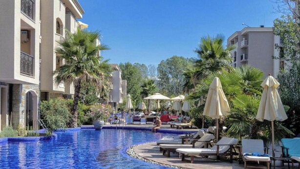 A resort pool area with lounge chairs and umbrellas, surrounded by palm trees and buildings. - Apartment Sleeps 4 with Pools Access - Smart TVs (Sunny beach)