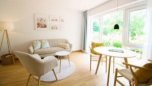 A modern living room with a sofa, chairs, a round table, and a large window with a view of greenery. - Vacation apartment small quiet oasis - forest edge location, central in Scharbeutz - House (Scharbeutz)