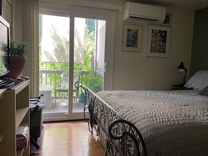 A bedroom with a bed, a desk with a computer, a chair, a potted plant, and a balcony with a view of greenery.