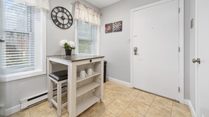 Une pièce avec une porte blanche, une horloge au mur, une fenêtre avec des stores, une petite table sur laquelle se trouve une plante, et une poubelle.