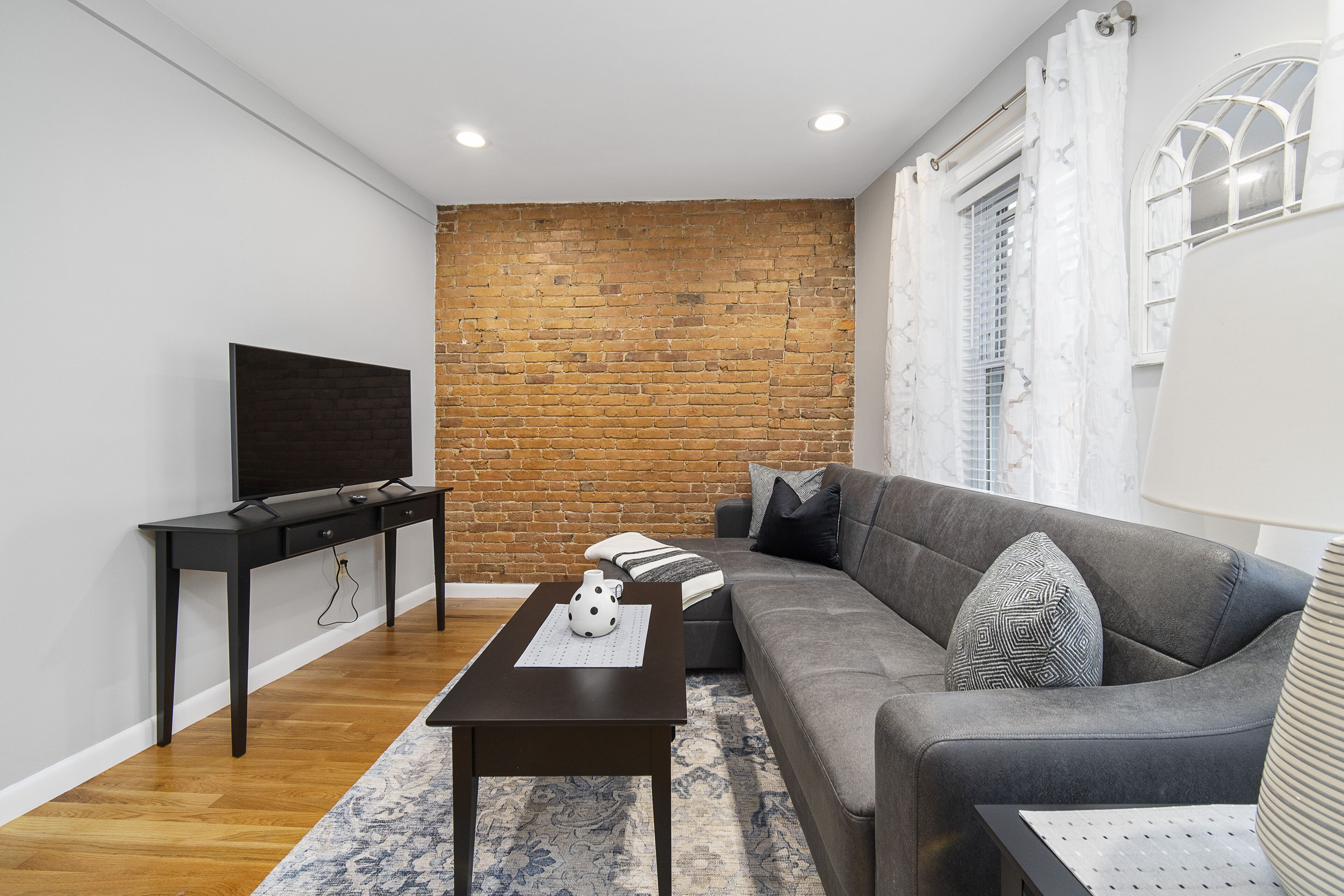 A modern living room with a brick wall, a grey sofa, a black TV stand, and a patterned rug.