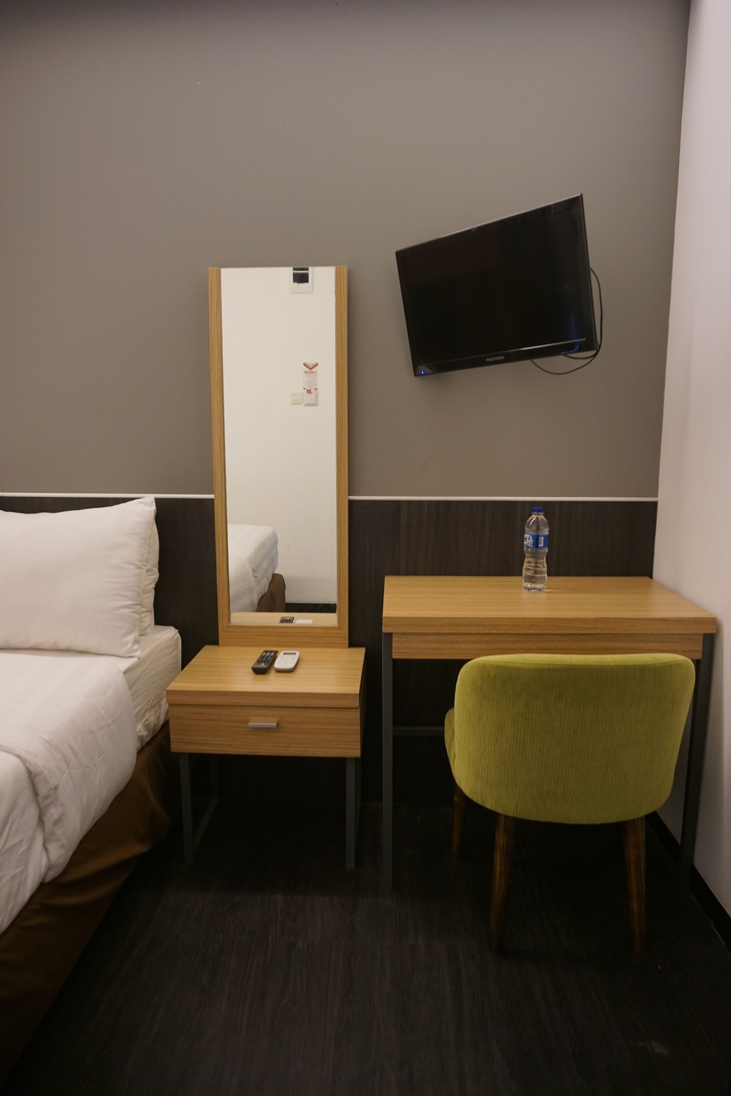 A hotel room with a bed, a desk with a chair, a flat-screen TV mounted on the wall, and a water bottle on the desk.