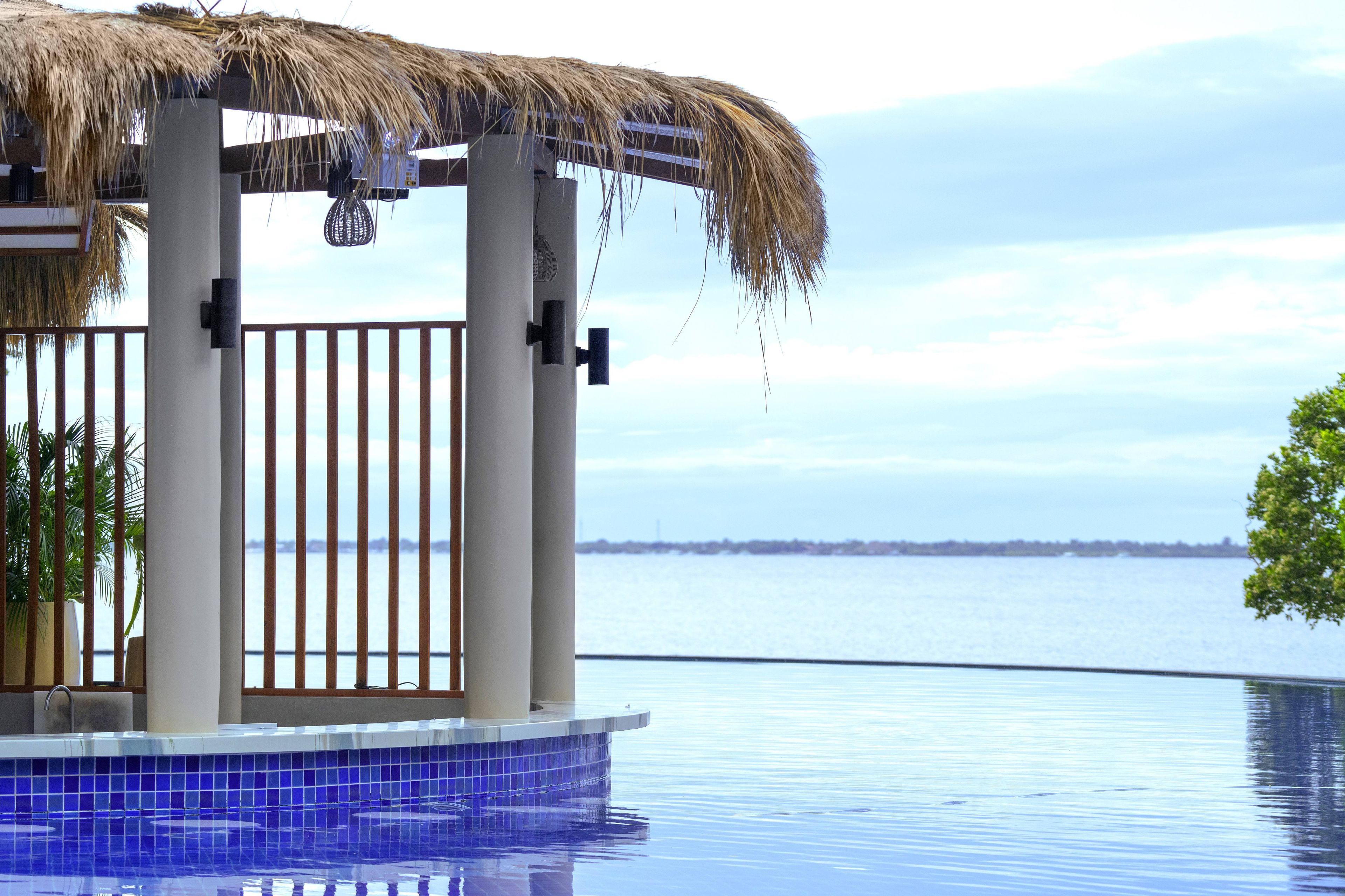 Un espace aménagé au bord de la piscine, doté d’une structure avec toit de chaume, offrant une vue sur l’eau et sur la côte, qui se dessine au loin.