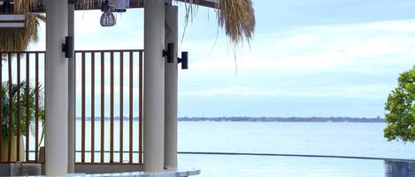 Un espace aménagé au bord de la piscine, doté d’une structure avec toit de chaume, offrant une vue sur l’eau et sur la côte, qui se dessine au loin.