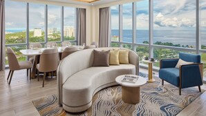 Una sala de estar moderna con un sofá curvo, una mesa de centro redonda y un comedor con sillas y una mesa. Amplias ventanas ofrecen vistas a la ciudad y al mar.