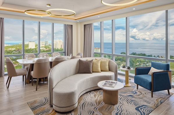 A modern living room with a curved sofa, a round coffee table, and a dining area with chairs and a table. Large windows offer a view of the city and the sea.