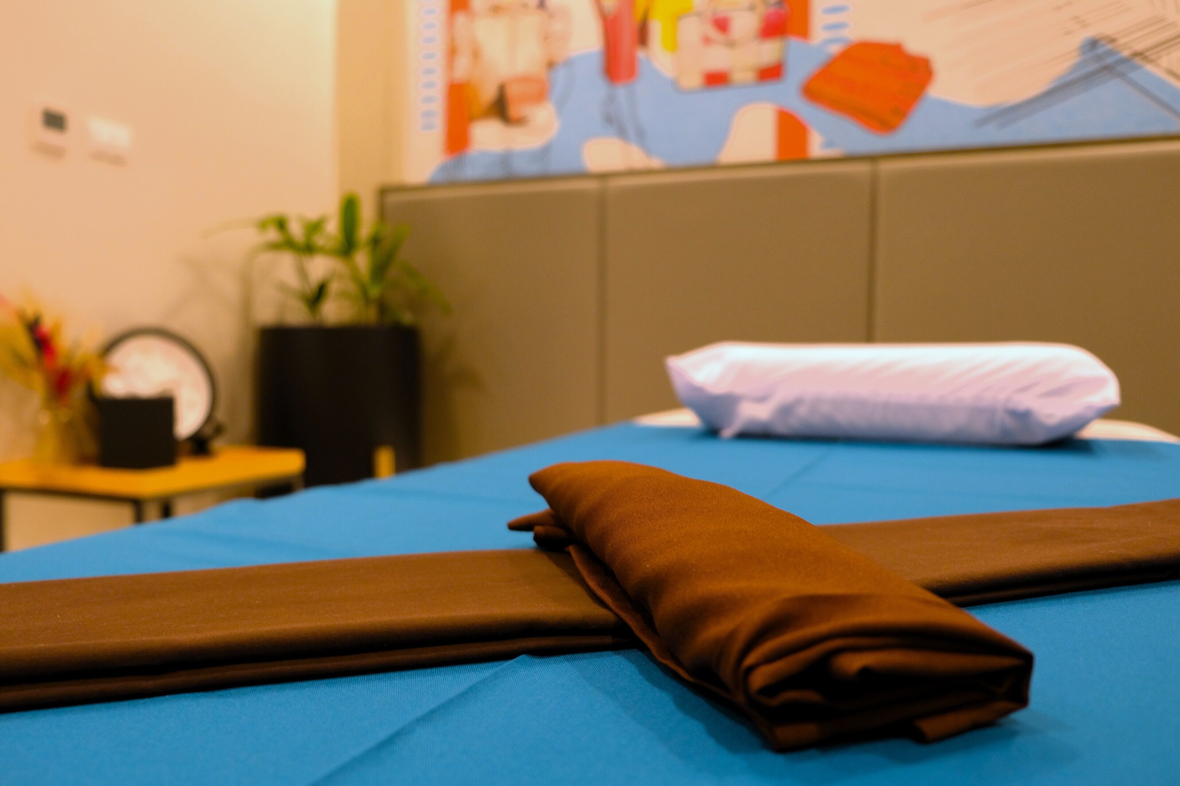 A neatly made bed with a blue bedspread and brown pillows, a small table with a clock and a plant, and a colorful wall mural in the background.