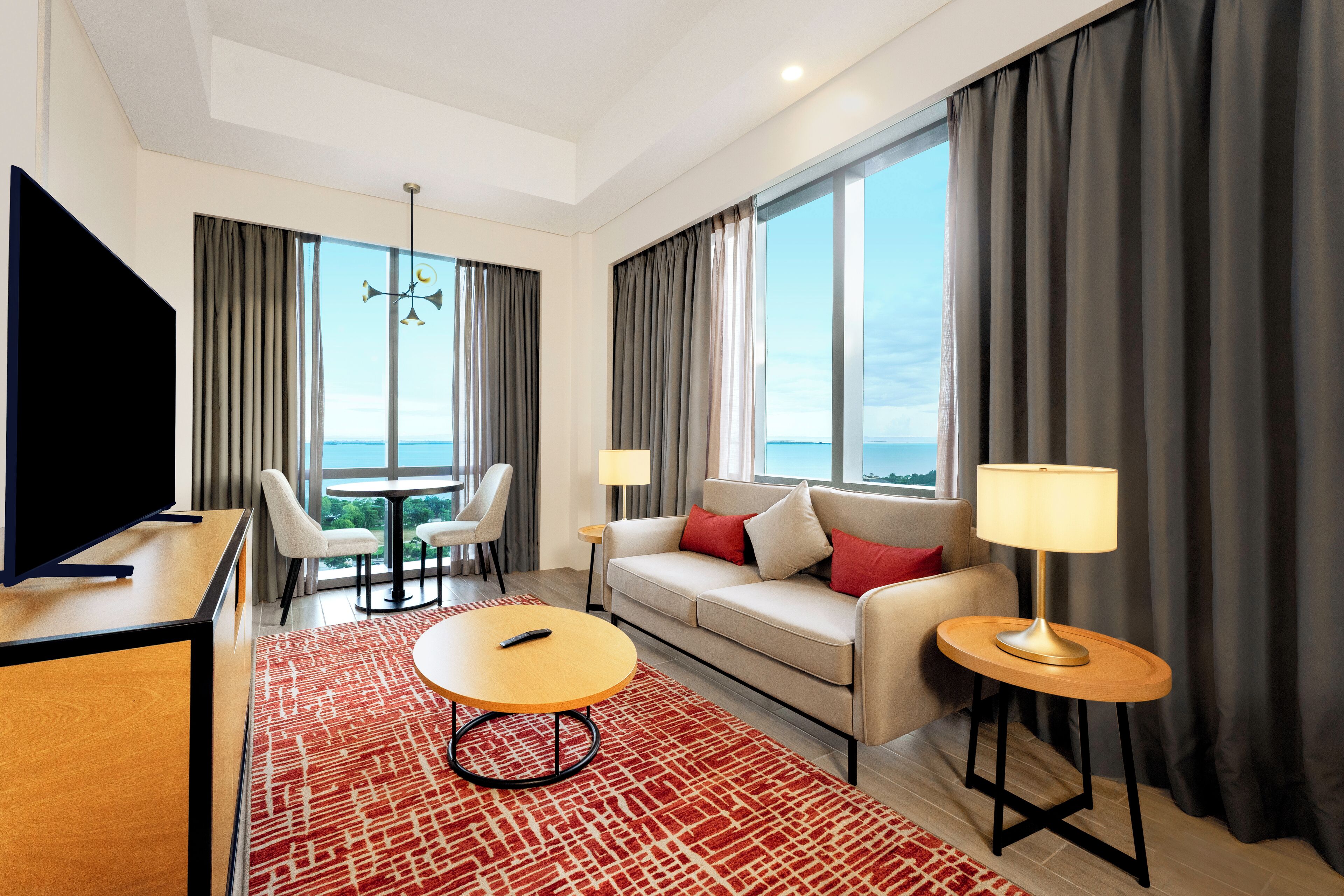 Une chambre d’hôtel moderne dotée d’une grande fenêtre, d’un canapé, d’une télévision, d’une table basse et d’une lampe.