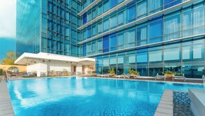 Un hotel moderno con una amplia piscina exterior, una zona de estar cubierta y un edificio con fachada de vidrio.