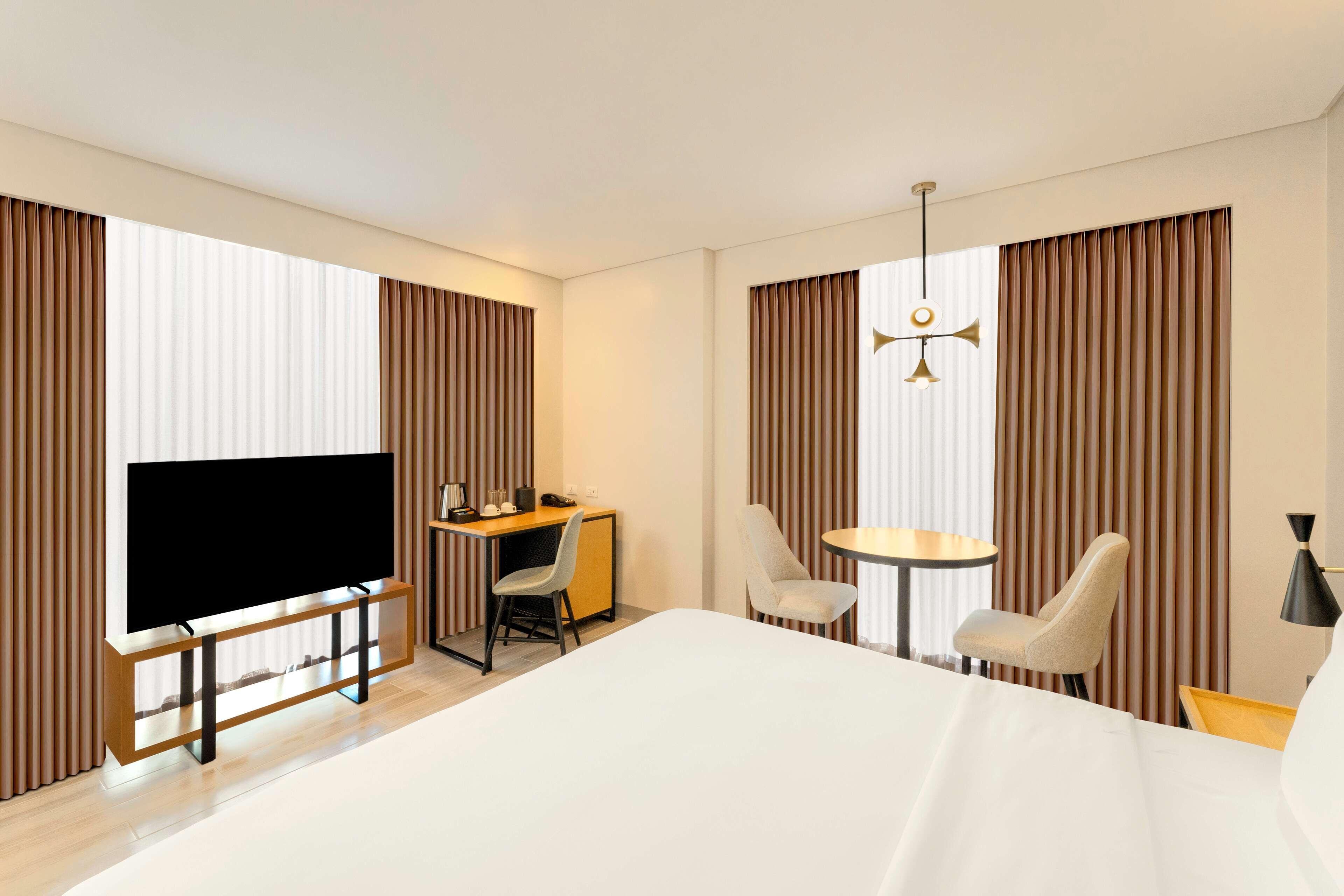 A hotel room with a large bed, a desk with a chair, a small round table with two chairs, a TV, and a ceiling light fixture.