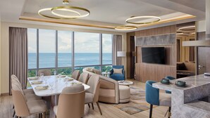 Une chambre d’hôtel moderne dotée d’un coin repas, d’un canapé, d’une télévision et offrant une vue sur la mer.