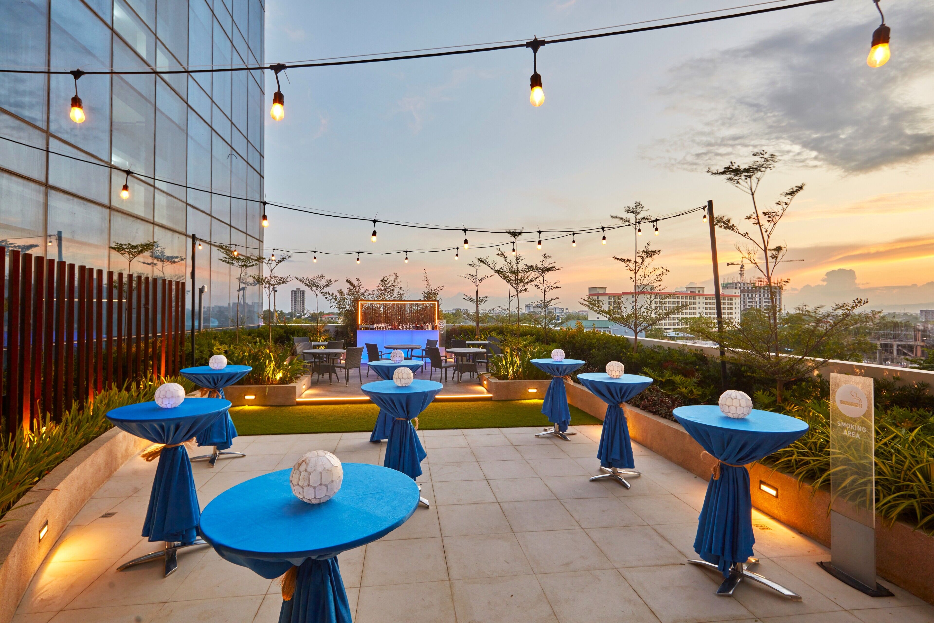 An outdoor seating area with tables and chairs, string lights, and a view of the city skyline at sunset.