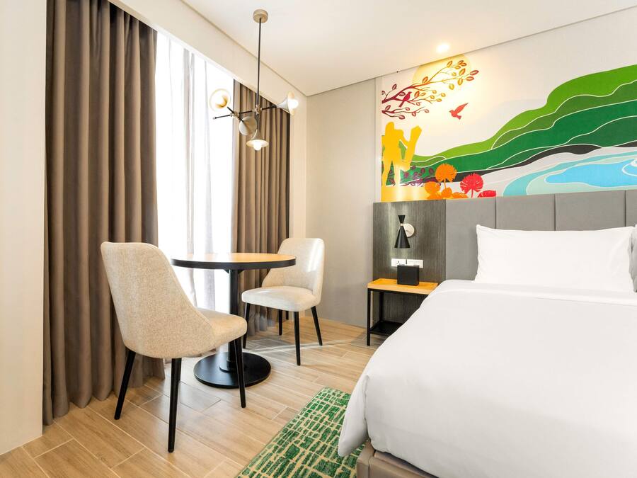 Een hotelkamer met een bed, een bureau met twee stoelen, een bijzettafeltje, een lamp en een groot raam met gordijnen.