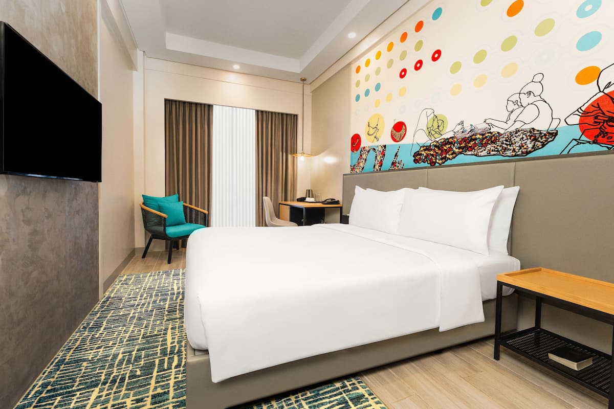 Hotelkamer met een groot bed, een aan de muur bevestigde televisie, een bureau met stoel, en een kleurrijke muurschildering.