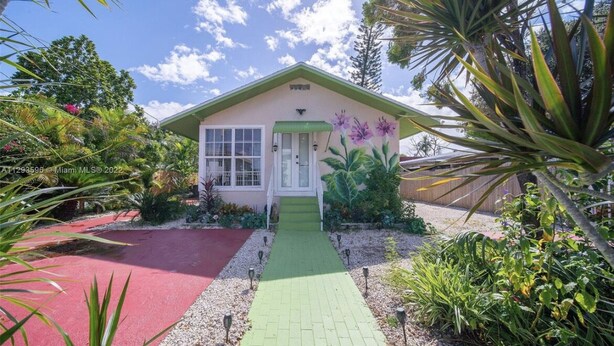 Une maison avec un chemin verdoyant menant à la porte d’entrée, entourée de plantes tropicales et bordée d’une zone rouge sur le côté gauche.