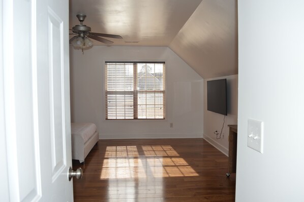 Una habitación con ventilador de techo, un ventanal con persianas, una cama y un televisor montado en la pared.