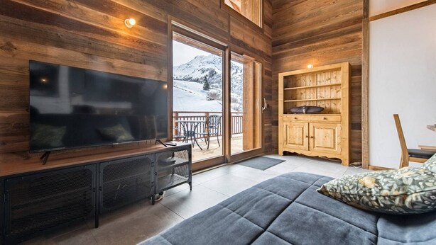 A cozy living room with a large flat-screen TV, wooden walls, a sofa, and a sliding glass door leading to a balcony.