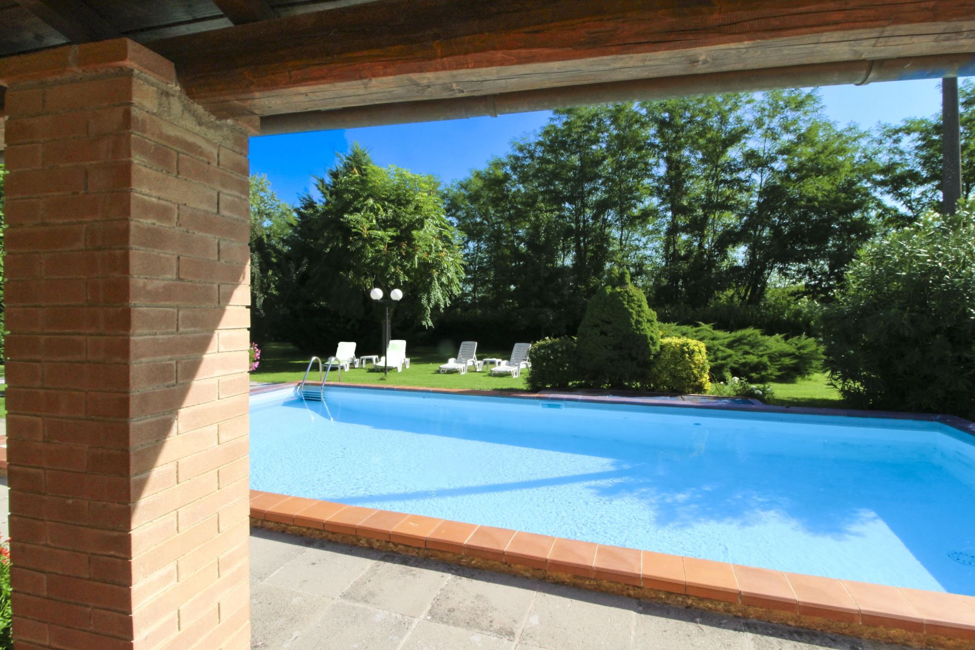 A swimming pool with a brick wall on one side and a view of trees and lawn chairs in the background.
