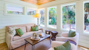 Un salón acogedor con un sofá color beige, una mesa de centro y un sillón blanco. Amplias ventanas ofrecen vistas a la vegetación exterior.