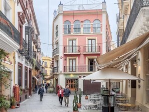 Een smalle straat met gebouwen aan beide kanten, waaronder een roze gebouw met balkons en een café met terras.