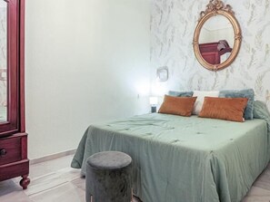 Een slaapkamer met een bed, een rond poefje, een dressoir en een spiegel aan de muur.