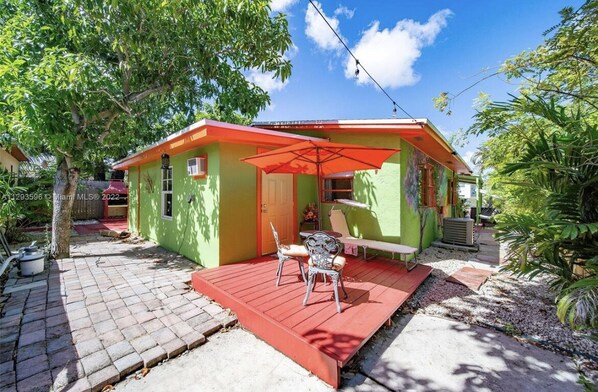 A small, green house with a red roof and a patio area with two chairs and a table.