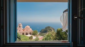 A window view of a coastal town with a prominent building featuring a tower, lush greenery, and the sea in the background. - Villa la Campanella in Ravello (Scala)