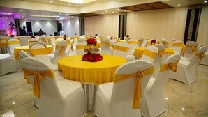 A banquet hall with round tables, white chairs with yellow sashes, and a central table with a yellow tablecloth and red flowers. - Comfort Hotel Amritsar (Amritsar)