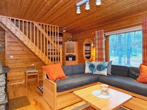 Interior de cabana com escada de madeira, um sofá, uma mesa de jantar e vista para a floresta pela janela.